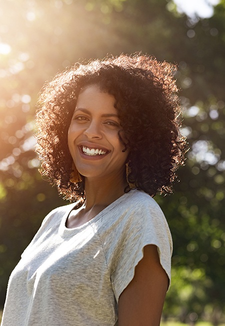 Woman in gray t-shirt outside smiling in front of a green tree